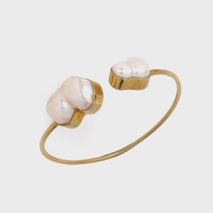 Puede incluir: Una pulsera abierta de metal fino en tono dorado con dos perlas barrocas blancas, irregulares y brillantes, engastadas en biseles dorados en cada extremo de la banda. Las perlas tienen una textura orgánica irregular y un brillo suave.