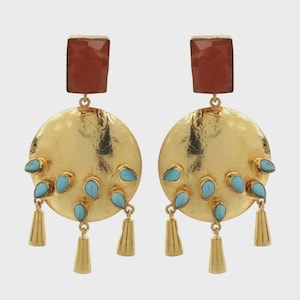 Op de afbeelding: Een paar goudkleurige oorbellen met een rechthoekige roodbruine steen aan de bovenkant, verbonden met een grote ronde gehamerde gouden schijf. De schijf is versierd met meerdere kleine turquoise druppelvormige stenen en drie bungelende gouden kegels.