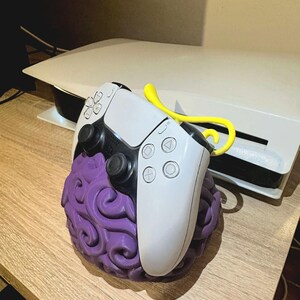May include: A white and black video game controller resting on a purple, textured, spherical stand with a swirling pattern. A bright yellow, curved, stem-like attachment extends from the top of the purple base behind the controller.