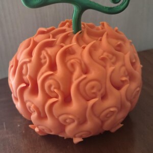 May include: A spherical orange object with a textured surface featuring swirling, flame-like patterns across its exterior. A green, branched stem with a curled tip extends from the top center of the object, which has a matte finish.