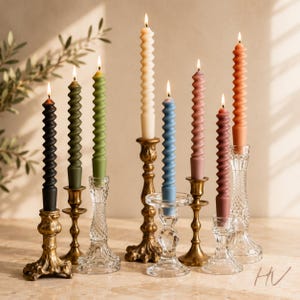 Puede incluir: Una colección de siete velas cónicas en espiral encendidas en colores negro, verde oliva, verde salvia, crema, azul, malva y terracota, sostenidas en varios candelabros ornamentados de latón y cristal tallado transparente.