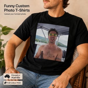 May include: A black t-shirt featuring a square graphic print of a person wearing bright yellow swimming goggles on a boat. The shirt includes text that reads Funny Custom Photo T-Shirts and Upload your funniest photo, with a Printed in Australia label.