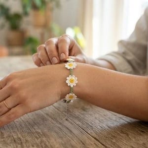 Puede incluir: Una pulsera que presenta una serie de motivos de flores de margarita blancas y amarillas conectados por un cordón verde fino. El diseño floral está elaborado con un intrincado trabajo de hilo y la pulsera incluye una cadena de metal.