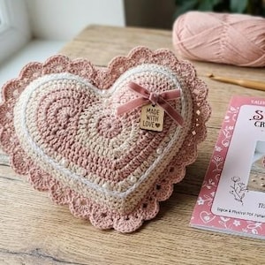 Op de afbeelding: Een hartvormig gehaakt kussen met een patroon van oudroze en crèmekleurig garen en een geschulpte rand. Het kussen is versierd met kleine witte kralen, een roze fluwelen strik en een houten label met de tekst Made With Love.