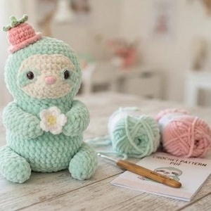 Può includere: Un peluche a forma di bradipo lavorato all'uncinetto color verde menta, con un piccolo cappello a forma di fragola rosa e un fiore bianco tra le zampe. Accanto ci sono due gomitoli di lana, un uncinetto in legno e un documento Crochet Pattern PDF.
