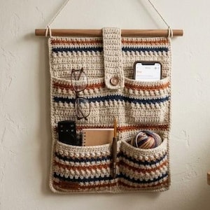 May include: A beige crochet wall organizer with horizontal stripes in navy blue and burnt orange. It features four pockets, a central tab with a wooden button, and a wooden dowel for hanging. The pockets contain eyeglasses, a remote, a notebook, a crochet hook, and yarn.