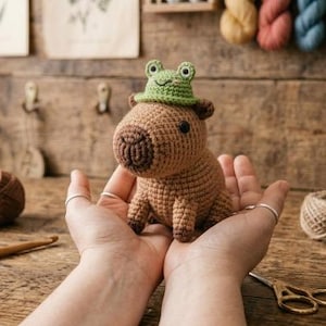 May include: A brown crocheted capybara figurine featuring a small green frog-shaped hat perched on its head. The capybara has a rounded body, dark brown stitched eyes, and a textured yarn surface. A separate green frog hat sits nearby.