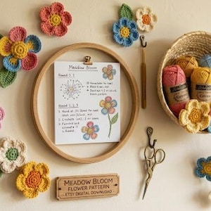 May include: A wooden embroidery hoop holding a Meadow Bloom pattern sheet, surrounded by various crocheted flowers in pink, yellow, blue, and green. A woven basket contains yarn skeins and more flowers, with scissors and a hook hanging on the wall.