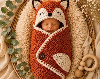 May include: A crochet baby swaddle blanket designed to look like a fox, featuring a hood with pointed ears, closed eyes, and a small black nose. The swaddle is primarily burnt orange with cream-colored accents and a single dark brown button closure.
