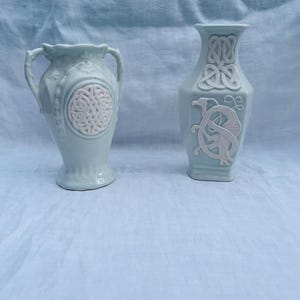 May include: Two pale blue ceramic vases with white relief details. The left vase features two handles and a circular Celtic knot design. The right vase has a hexagonal shape with a top Celtic knot pattern and a lower relief of a bird figure.