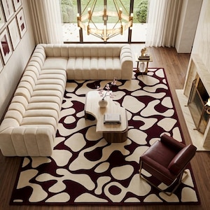 May include: A large area rug featuring a bold, irregular cream and deep burgundy organic pattern. The rug is positioned beneath a cream-colored sectional sofa, a curved wooden coffee table, and a dark red leather armchair on a wooden floor.