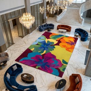 May include: A large rectangular area rug featuring a vibrant, oversized floral pattern in bold shades of magenta, bright yellow, orange, lime green, and deep blue, centered in a spacious lobby with marble flooring and curved seating arrangements.