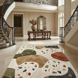 May include: A rectangular area rug featuring a cream-colored base with large, abstract, organic shapes in shades of olive green, tan, rust orange, and charcoal gray. Some sections contain a speckled terrazzo-inspired pattern with multi-colored chips.