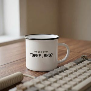 Puede incluir: Una taza de esmalte blanco con borde y asa negros, que presenta el texto impreso en negro "Do you even TOPRE, BRO?" en el frente. La taza tiene un cuerpo cilíndrico y un acabado liso, colocada junto a un teclado mecánico sobre una superficie de madera.