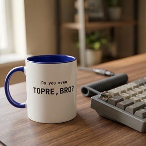 Puede incluir: Una taza de cerámica blanca con interior y asa de color azul. El frente de la taza presenta el texto impreso en negro "Do you even TOPRE, BRO?" en una fuente sans-serif clara. La taza tiene un cuerpo cilíndrico y un asa curva en el lateral.