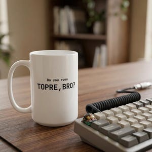 Puede incluir: Una taza de cerámica blanca con un asa en el lado izquierdo, que presenta el texto impreso en negro "Do you even TOPRE, BRO?" centrado en la parte frontal. La taza está colocada junto a un teclado mecánico con teclas grises y blancas.