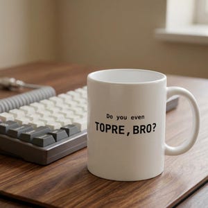 Puede incluir: Una taza de cerámica blanca con el texto impreso en negro 'Do you even TOPRE, BRO?' en la parte frontal. La taza tiene una forma cilíndrica estándar con un asa curva en el lado derecho, situada junto a un teclado mecánico sobre una superficie de madera.