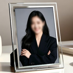 Puede incluir: Un marco de fotos rectangular de metal plateado con un diseño de doble borde. El marco contiene el retrato de una persona con cabello largo y oscuro que viste un blazer negro. El texto SereneCanvasShop está impreso en blanco en la base.