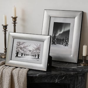 May include: Two silver-toned picture frames with wide, beveled borders. The horizontal frame displays a black and white photograph of a tree-lined path, while the vertical frame shows a black and white photograph of a snowy forest with aurora borealis.