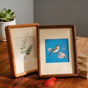 May include: Two rectangular wooden picture frames with cream-colored matting. The left frame displays a green eucalyptus branch illustration. The right frame features a small bird perched on a flowering branch against a bright blue background.