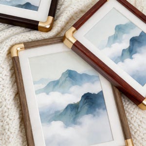 May include: Two rectangular picture frames with dark wood grain borders and polished gold-tone metal corner accents. Each frame contains a watercolour-style print depicting blue mountain peaks rising above white clouds with a white mat border.