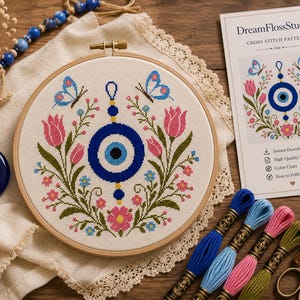 May include: A cross stitch pattern in a wooden hoop featuring a central blue evil eye symbol surrounded by pink tulips, green foliage, and two blue butterflies. Beside it is a printed instruction card labeled DreamFlossStudio, embroidery floss, and scissors.