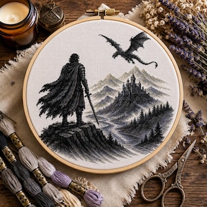 May include: A circular wooden embroidery hoop containing a cross-stitch design of a cloaked figure holding a sword, standing on a cliff overlooking a castle, mountains, and a flying dragon. The scene is rendered in shades of black, gray, and white.