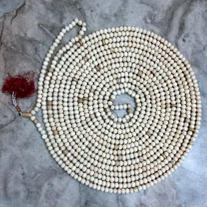 May include: A long strand of off-white spherical beads arranged in a spiral pattern. The beads are primarily cream-coloured with occasional light brown accent beads interspersed throughout. A deep red tassel is attached to one end of the bead strand.