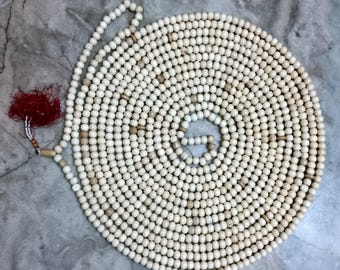 May include: A long strand of off-white spherical beads arranged in a spiral pattern. The beads are primarily cream-coloured with occasional light brown accent beads interspersed throughout. A deep red tassel is attached to one end of the bead strand.