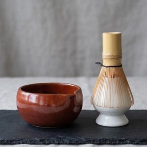 May include: A glossy, deep reddish-brown ceramic bowl with a small pouring spout sits beside a bamboo matcha whisk resting on a white ceramic stand. The whisk features thin, light-coloured bamboo tines tied with a thin black cord.
