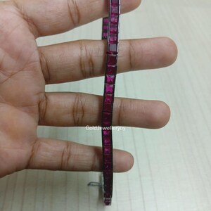 May include: A thin, flexible bracelet featuring a continuous row of square-cut, vibrant magenta-pink stones set in a dark metal frame. The piece includes a small metal clasp at the bottom and displays the text GoldJewellery05 in the centre.