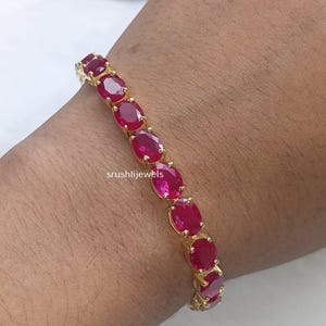 May include: A gold-toned bracelet featuring a series of oval-cut, vibrant pinkish-red gemstones set in individual metal prongs. The jewellery piece displays the text srushtijewels in small white lettering across the centre of the bracelet design.