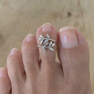 May include: A silver-toned toe ring featuring a delicate, open-ended design shaped like two overlapping leafy branches. The metal band wraps around the toe, with the leaf motifs extending outward to create a botanical appearance on the skin.