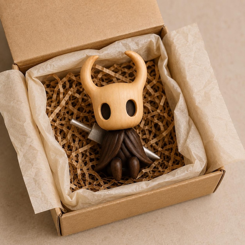 May include: A wooden figurine of a character with a pale, smooth, rounded head featuring two large, dark, oval-shaped eye sockets and two curved horns, wearing a dark brown, textured cloak and holding a silver-colored sword in a box.
