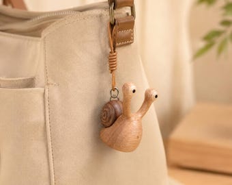 May include: A light beige canvas shoulder bag with a wooden snail charm attached to the strap. The charm features a smooth, light-toned wooden body with two eye stalks and a darker, spiraled wooden shell, suspended by a thin leather cord.