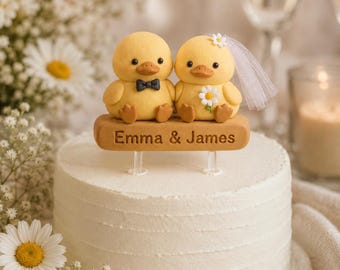 May include: A cake topper featuring two yellow ducklings sitting on a tan rectangular base with the text Emma & James. One duckling wears a black bow tie, and the other wears a white daisy and a sheer white veil. The topper sits on a white cake.