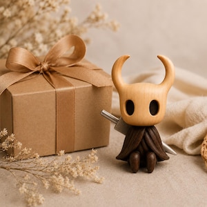 May include: A small wooden figurine of a character with a pale, smooth, horned head and large dark oval eyes. The figure has a dark brown, textured cloak and holds a silver-colored sword behind its back, positioned next to a brown gift box.