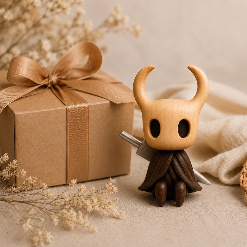 May include: A small wooden figurine of a character with a pale, smooth, horned head and large dark oval eyes. The figure has a dark brown, textured cloak and holds a silver-colored sword behind its back, positioned next to a brown gift box.