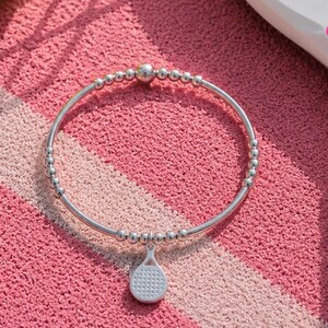 May include: A silver-toned stretch bracelet featuring a series of small metallic beads and one larger central bead. A small, flat, silver-toned charm shaped like a pickleball paddle with a textured grid pattern hangs from the bottom of the bracelet.