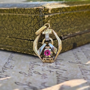 Op de afbeelding: Een gouden hanger met een geometrisch frame en een centrale ronde roze edelsteen. Het ontwerp bevat een klein zilverkleurig accentstuk met een minuscuul helder steentje boven de roze edelsteen en een gouden hanger aan de bovenzijde.