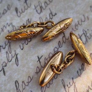 Peut inclure: Une paire de boutons de manchette en métal doré de forme ovale allongée avec des détails floraux gravés. Le dos de l'un des boutons porte clairement l'inscription FIX. Ces accessoires utilisent un mécanisme à maillons pour relier les deux extrémités.