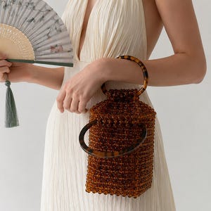 May include: A structured handbag crafted from translucent amber-colored beads in a square silhouette, featuring two circular tortoiseshell-patterned handles. The bag is paired with a folding hand fan with a light gray fabric and a green tassel.
