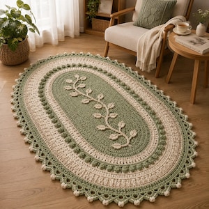 May include: An oval crochet rug featuring a sage green center with a raised white vine and leaf pattern. The rug has concentric borders in cream and sage green, finished with a scalloped edge and small white bobble accents along the perimeter.