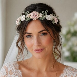 Puede incluir: Una diadema floral con flores tejidas a ganchillo en tonos rosa suave, crema y blanco, acentuada con hojas tejidas en verde salvia y pequeñas perlas blancas en el centro de cada flor, dispuestas en un diseño de banda decorativa continua.