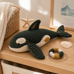 May include: A dark green and cream crocheted orca toy featuring a small dorsal fin with yellow stitching, a rounded tail, and a matching miniature crocheted life ring with a thin twine string resting beside it on a wooden surface.