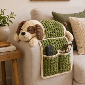 May include: A crochet dog-shaped sofa armrest organizer in cream and dark brown with a green textured body. The organizer features two front pockets containing a black remote control and brown-rimmed glasses, draped over a light-colored sofa arm.
