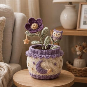 Peut inclure: Un cache-pot au crochet crème et violet orné d'un motif de croissant de lune et d'étoiles. À l'intérieur se trouve une fleur violette au crochet avec un cœur beige, une petite étoile suspendue et un petit hibou violet aux yeux fermés.
