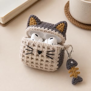 Puede incluir: Una funda de ganchillo con forma de gato para auriculares inalámbricos, con un frente beige de textura de gofre, orejas gris oscuro con detalles amarillos y bigotes negros bordados. Un pequeño dije de espina de pescado en gris y amarillo cuelga.