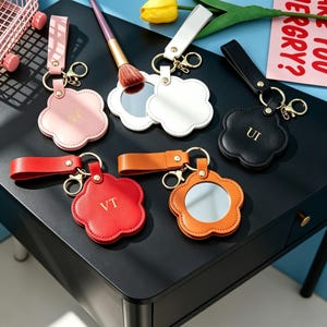 May include: A collection of flower-shaped keychains with attached mirrors, featuring leather-like straps and gold-tone hardware. The keychains come in pink, white, black, red, and orange, with some displaying gold-embossed initials like SH, UI, and VT.