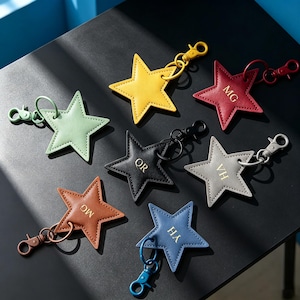 May include: A collection of seven star-shaped keychains in yellow, red, light green, black, grey, brown, and blue. Each keychain features a metal lobster clasp and ring. Some stars are embossed with gold-colored initials including MG, QR, VH, and YH.
